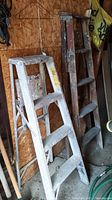 Photo showing side by side both metal 4-foot ladder and wooden 5-foot ladder leaning against wall with paint and wear marks.