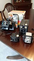 Photo showing five vintage cameras: Polaroid Land Camera, Super Shooter, Brownie Target Six-16, Brownie Starflex, Baby Brownie Special. The Polaroid camera box is also present.