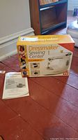 Photo of boxed Dressmaker Sewing Center Model 998B and instruction manual on hardwood floor, showing box details and manual front page.