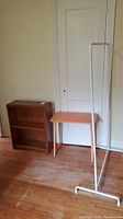 Small wooden bookshelf, painted table, and white metal clothes rack positioned against a white door on hardwood floor