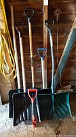 Five snow shovels and one metal extender rod standing upright against a garage wall on a concrete floor.