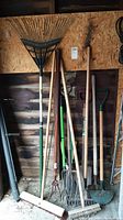 Full view of the garden tools leaning against a wall including rake, pitchforks, brooms, clippers, and spade.