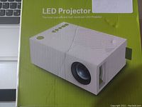 Photo of the white LED projector in its box showing front lens and top surface with text.
