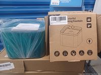 Photo of the pet drinking fountain in teal plastic alongside its retail box showing product description and features.