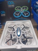 Photo of the Q9s remote control drone in white and blue with black propellers, inside its original styrofoam packaging in front of a black box with product details.