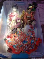 Plastic bin containing multiple Asian dolls in various colored traditional costumes, one wrapped in plastic and a decorative fan visible