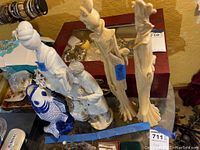 Photo showing a blue and white ceramic fish, man playing flute figurine, and two tall carved cream-colored Asian figurines, displayed on a glass surface with wood box in background.