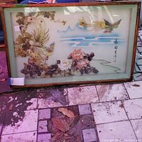 Full view of framed Asian floral bird artwork resting on stone tiles outdoors; artwork features birds and flowers with Asian characters in bottom right.