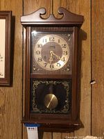 Front view of Centurion wall clock showing full wooden case, clock face with gold numerals and pendulum behind glass door.