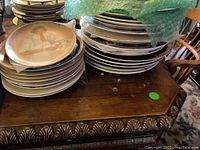 Overall view of stacks of collectible plates on a wooden surface including some with gold edges and decorative art.