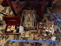 Overview of multiple small boxes and trays containing assorted costume jewelry laid on floral carpet beneath wooden table