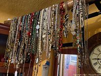 Photo showing a variety of costume jewelry necklaces hanging on a wooden railing with a wall clock in the background.
