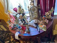 Photo showing multiple Disney and other figurines on a wooden table with clocks in the background. Items include large Mickey Mouse and Disney princess figures.