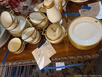 Assortment of white porcelain pieces with gold trim including tea pot, sugar bowl, creamer, cups, saucers and plates on wooden table