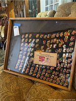 Framed Olympic pin collection of international flags in wooden frame, photographed on a patterned couch, visible reflections on glass, with an auction lot tag on the frame.