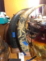 Pair of carved bull horns, one with dragon design, standing upright on a wooden base on a table surrounded by other antiques and dishware.