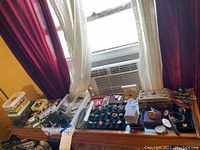 Tabletop near window showing multiple trays and boxes of watches, rings and bracelets spread across wood surface.