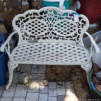 Front view showing the white wrought iron bench with lattice design and ornamental scrollwork in backrest and arms, placed on tiled floor with some outdoor items around.