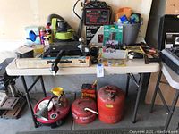 Full lot view showing variety of automotive items including battery charger, gas cans, cleaning supplies, tow hitch, and others on table and floor.