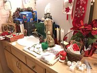 Wide view showing assorted Christmas decor, linens, small boxes, lights and figurines arranged on wooden cabinet surface.