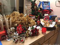 Wide view showing gold tinsel, poinsettia flowers, ceramic candle holders shaped like houses, electric candlestick lights, and assorted holiday decorations spread on a counter.