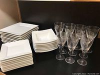 Stack of white square porcelain plates and bowls next to a set of crystal stemware glasses showing clear etched Flight pattern.