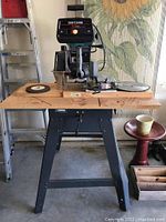 Full view of the Craftsman Table Saw mounted on the wooden bench with metal stand in a garage setting; various saw blades and tools on the bench.