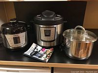 Two kitchen appliances sitting side by side: Instant Pot IP-LUX pressure cooker with steel body and digital display, and Cosori pressure rice cooker with black and stainless steel finish, plus recipe booklets and a stainless steel pot lid.