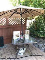 Patio table with Treasure Garden umbrella standing through the center, glass top table with metal frame, objects on the table include two tea light lantern boxes and other boxed items