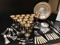 Overview of silver-plated stemmed goblets, various small spoons, flatware with pearlized handles, monogrammed plate, and shakers arranged on black surface.