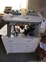 Woodcraft router table on top of white three-drawer cabinet with manuals and bits visible