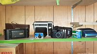 Four cameras including Polaroid, Nikon Oneshot 35mm, Kodak Instant, and Kodak Brownie models along with Fujifilm film and a film case on a green shelf against wood panel wall.