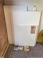 Four white canvasses leaning against wall on carpet, with various paint tubes laid in front on carpet