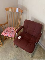 Two chairs side by side. On the left a wooden rocking chair with vertical slat back and pink seat cushion. On the right a metal framed upholstered arm chair with maroon fabric and padded armrests.