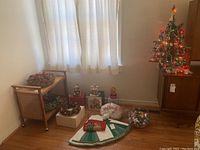 Overall view showing small artificial Christmas tree on a cabinet, assorted wreaths on a cart and on the floor, tree skirt, music boxes, glass bowl of ornaments, and other decorations grouped near a window.