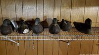 Six wooden vintage decoy ducks on a white wire shelf against wood-paneled wall, showing front and side views.