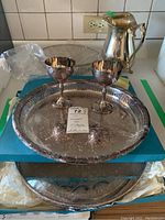 Full set displayed on a surface including the silver plated tray with intricate engraving, two goblets placed on tray, pitcher, and wire basket visible on side.
