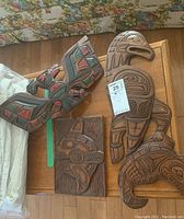 Three carved Indigenous wooden art pieces on a wooden surface: large signed wall plaque, painted Thunderbird carving, smaller relief plaque.