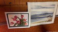 Two framed original watercolour paintings leaning against wood panel wall on carpeted floor, one floral and one landscape.