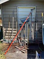 Photo showing a collection of garden tools including hedge trimmer, rakes, shovels, hoes, and small hand tools leaning against the side of a building outdoors.