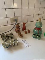 Wide view of multiple saki bottles and cups arranged on a white countertop with tiled backsplash, showing variety in size, color, and style of saki sets.
