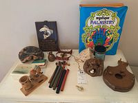 Photo showing a variety of decorative items on a white surface including a palmistry book, candle holder, wooden plaques, carved figures, string instrument, shell, bear figure, and colored sticks.