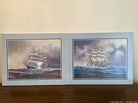 Two laminated nautical pictures side by side on wooden table showing white sailing ship schooners on ocean waves with cloudy skies, each approximately 20x10 inches