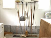 Photo shows all 9 tools standing upright against a wall in a room, including brooms, shovel, rake, hoe, and squeegee.