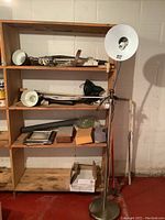 Wooden shelving unit containing various lamps, lighting parts, picture frames, and a metal floor lamp with a white round shade next to it.