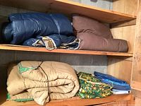 Shelf holding four pieces: blue and brown sleeping bags on upper shelf; tan blanket or tarp, green paisley tarpaulins, and blue tarp/fabric on lower shelf.