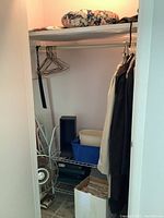 Closet space showing hanging women's XL coats including a light beige overcoat and a black coat, plus a blue CD storage box, white plastic bins, and a metal 3-speed table fan on bottom shelf.