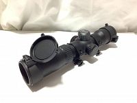 Side angled photo of black Excalibur TACT-100 tactical riflescope with lens covers closed, showing adjustment knobs.