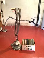 Photo showing a full view of the Holcomatic Mark XII floor buffer standing on a red floor next to a box filled with multiple round polishing pads and brushes.