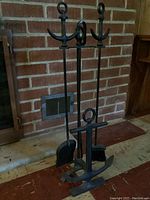 Photo showing full fireplace tool set with black cast iron anchor-themed stand and three tools in front of brick fireplace.